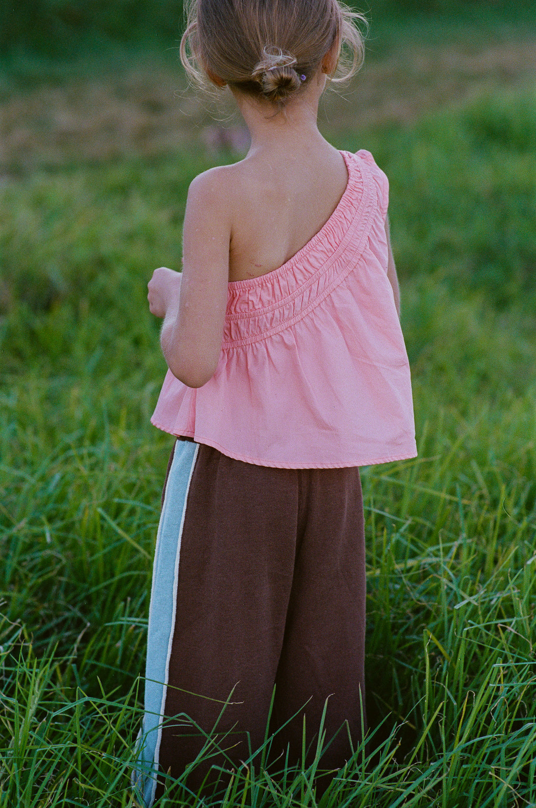girl wears one shoulder pink top in 100% cotton poplin with gathering detail and linen/cotton blend chocolate knit pants with blue stripe and piping