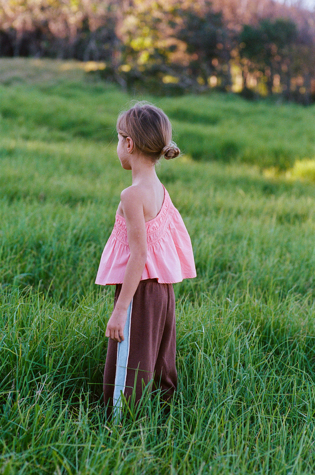 girl wears one shoulder pink top in 100% cotton poplin with gathering detail and linen/cotton blend chocolate knit pants with blue stripe and piping