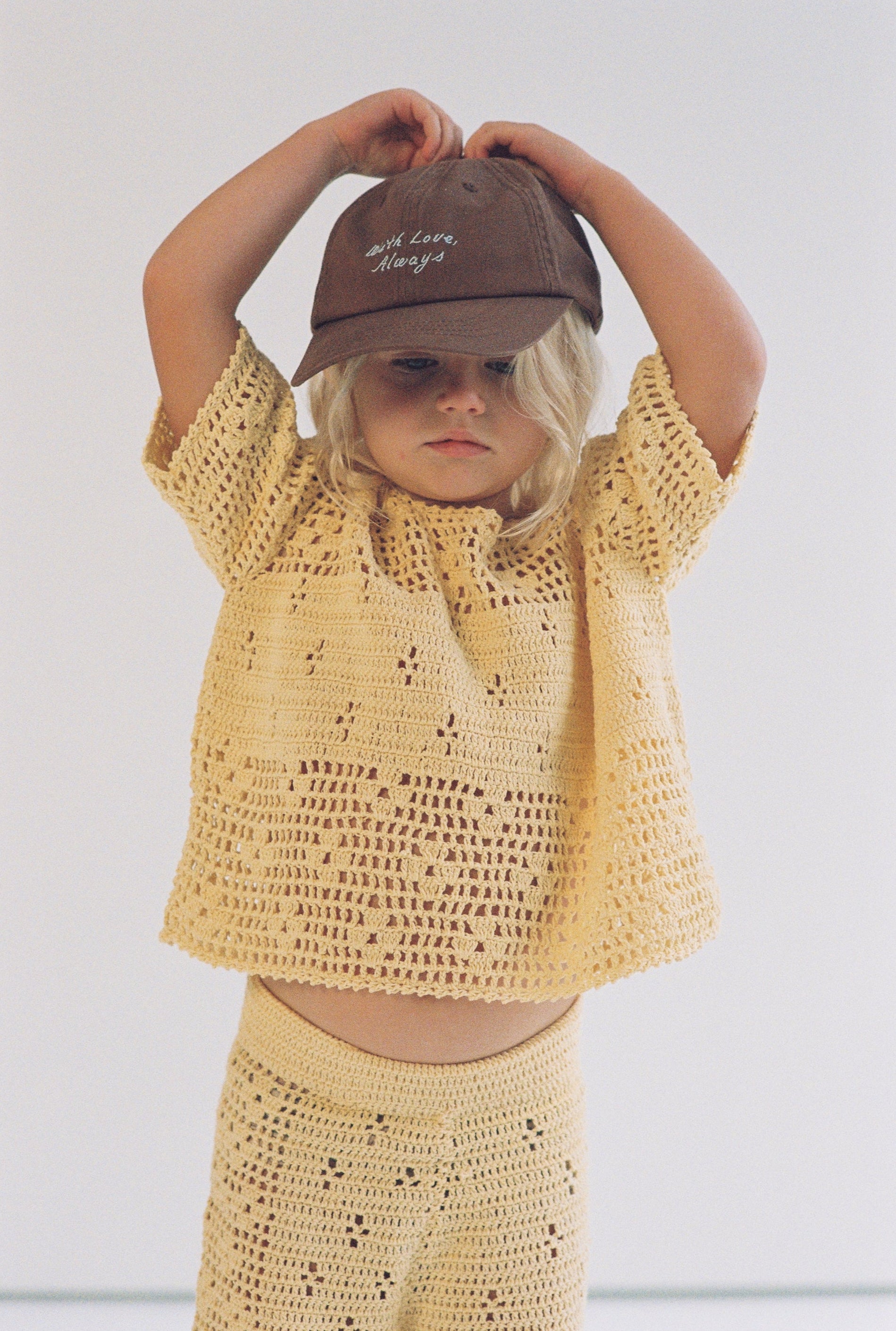 Child wearing a yellow crochet outfit and brown cap against a plain background