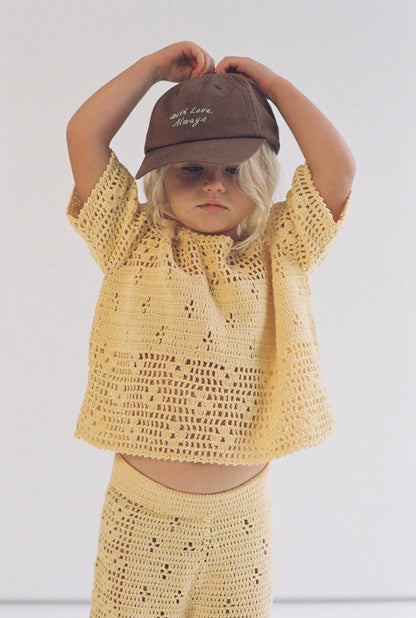 Child wearing a yellow crochet outfit and brown cap against a plain background